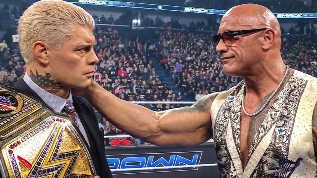 Two professional wrestlers, The Rock and Cody Rhodes, stand face-to-face in the wrestling ring, with a cheering crowd visible in the background.